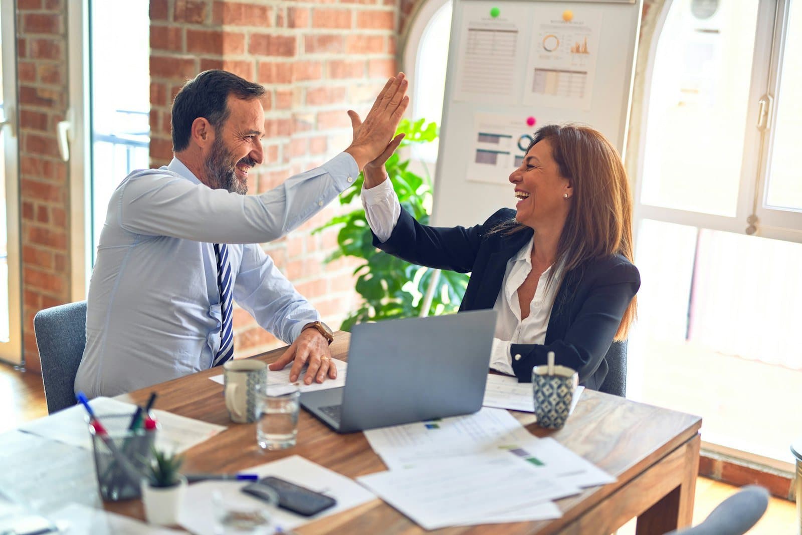 Two senior colleagues high-five across a desk covered in papers